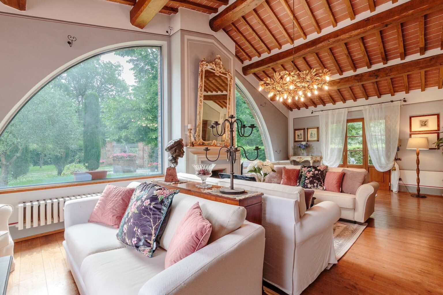 Cozy living room featuring a large arched window, wooden ceiling beams, and elegant decor. The space includes a comfortable white sofa adorned with colorful cushions and a stylish chandelier, creating a warm and inviting atmosphere. | Only on SopranoVillas