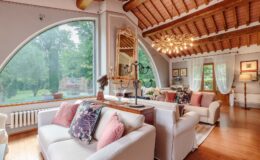 Cozy living room featuring a large arched window, wooden ceiling beams, and elegant decor. The space includes a comfortable white sofa adorned with colorful cushions and a stylish chandelier, creating a warm and inviting atmosphere.