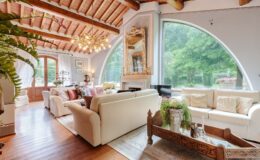 Spacious living room with high wooden ceiling, large windows overlooking greenery, elegant furniture, and decorative accents, creating a warm and inviting atmosphere.