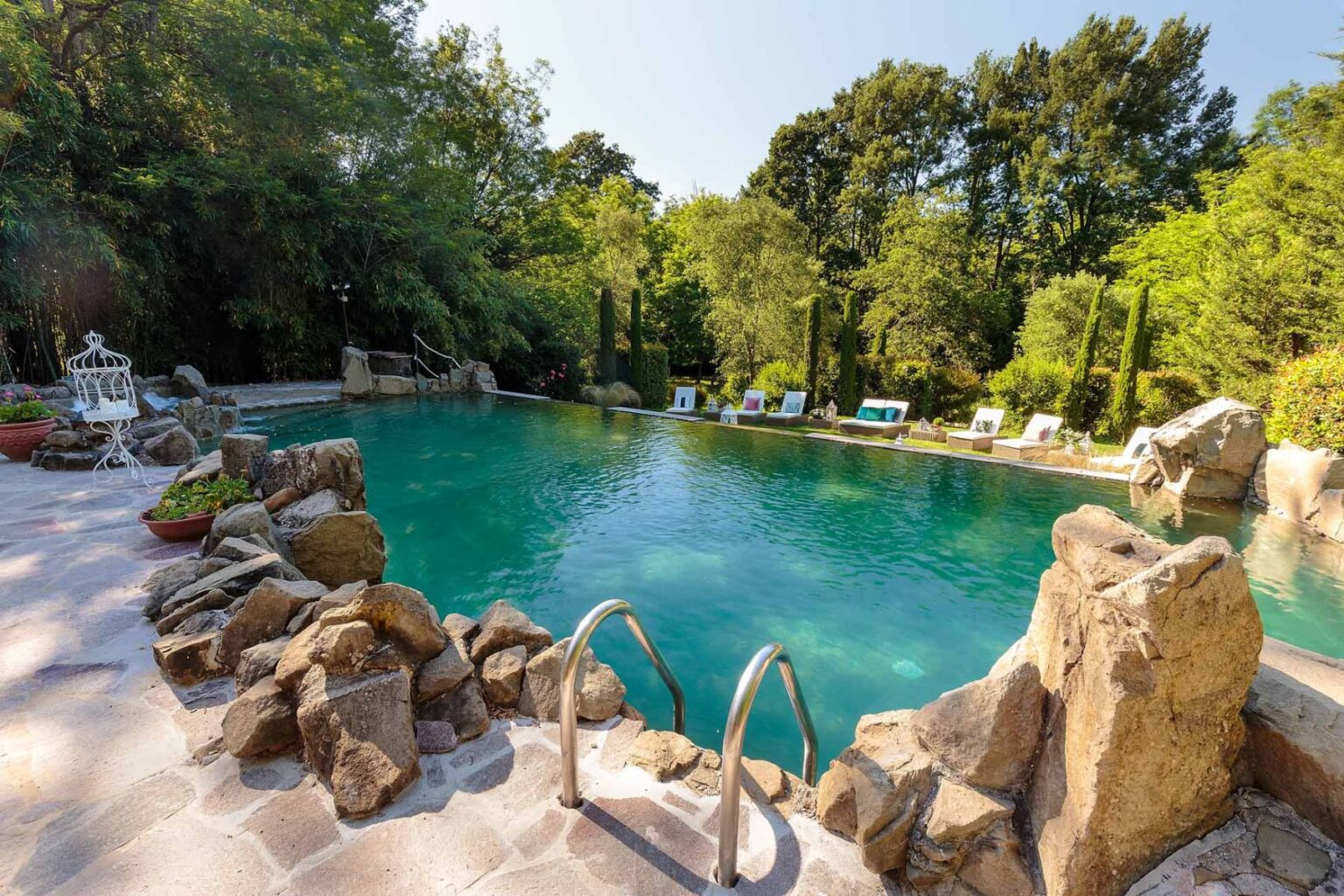 Scenic outdoor swimming pool surrounded by lush greenery and natural rock formations, featuring lounge chairs for relaxation and a clear blue water surface reflecting the sunlight. | Only on SopranoVillas