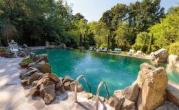 Scenic outdoor swimming pool surrounded by lush greenery and natural rock formations, featuring lounge chairs for relaxation and a clear blue water surface reflecting the sunlight.