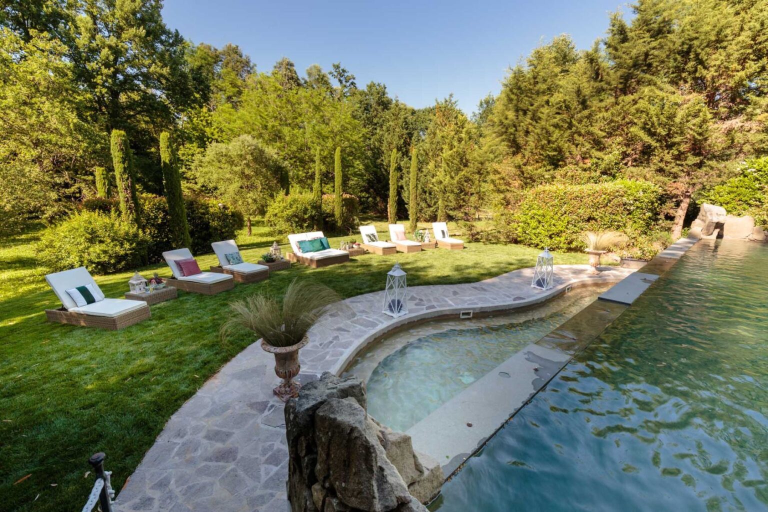 Lush outdoor pool area surrounded by greenery, featuring a curved pool, stone deck, and comfortable lounge chairs for relaxation. | Only on SopranoVillas