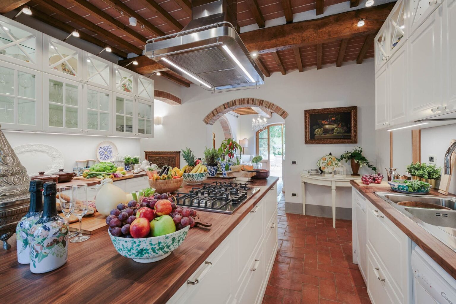 Spacious modern kitchen featuring a central island with a gas stove, wooden beams, and elegant cabinetry, adorned with fresh fruits and vegetables, creating a warm and inviting atmosphere. | Only on SopranoVillas