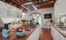 Spacious modern kitchen featuring a central island with a gas stove, wooden beams, and elegant cabinetry, adorned with fresh fruits and vegetables, creating a warm and inviting atmosphere.