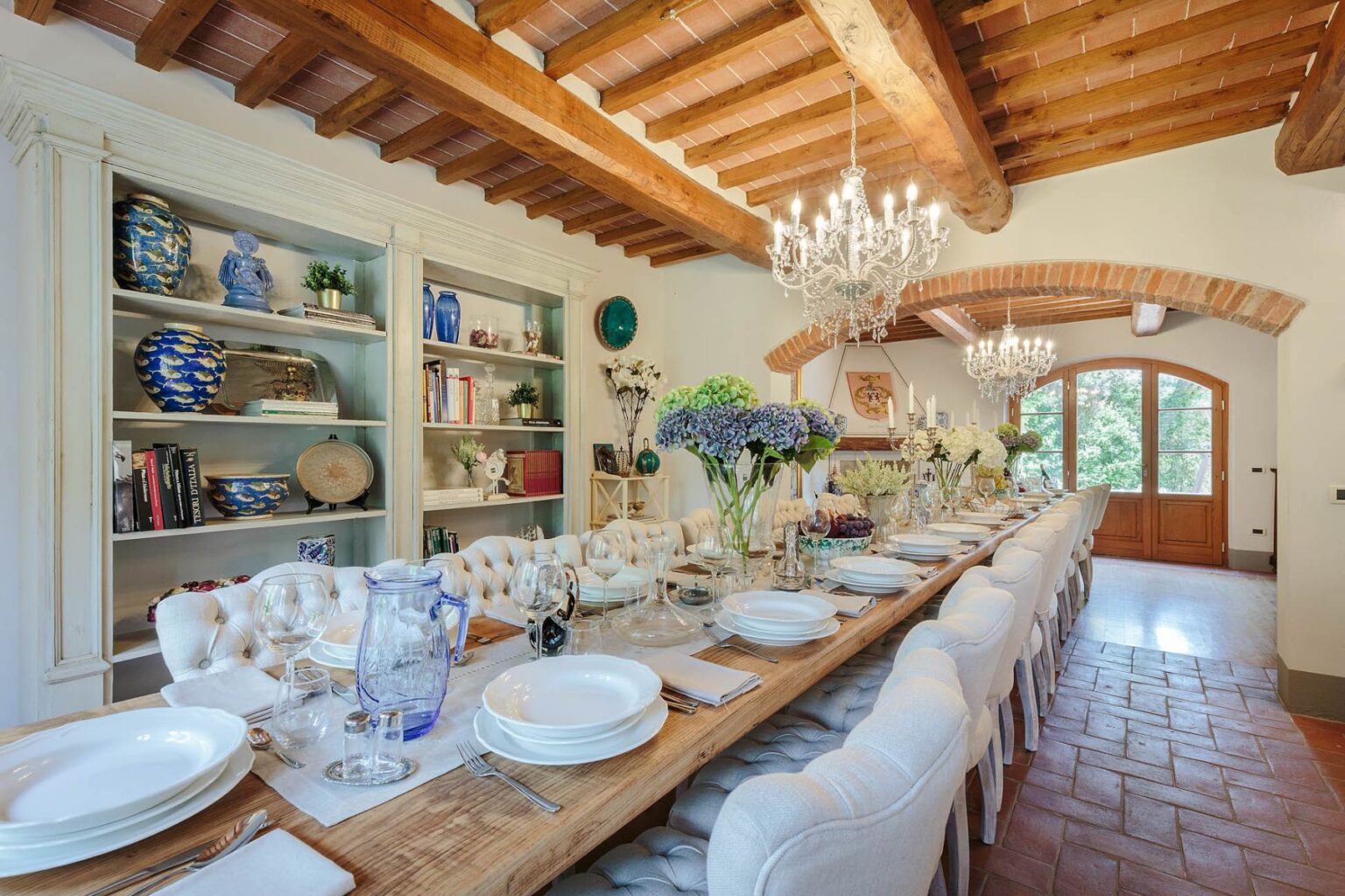 Elegant dining room featuring a long wooden table set for a meal, adorned with white plates and floral centerpieces. The space showcases rustic wooden beams, a stylish chandelier, and shelves filled with decorative items, creating a warm and inviting atmosphere. Large windows provide natural light and a view of the outdoors. | Only on SopranoVillas