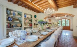 Elegant dining room featuring a long wooden table set for a meal, adorned with white plates and floral centerpieces. The space showcases rustic wooden beams, a stylish chandelier, and shelves filled with decorative items, creating a warm and inviting atmosphere. Large windows provide natural light and a view of the outdoors.