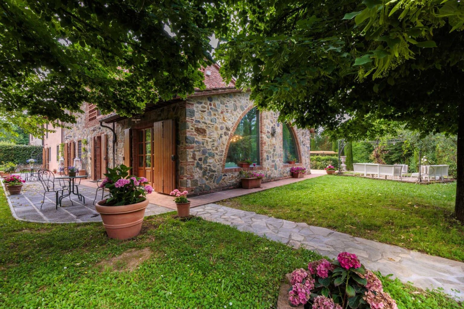 Charming stone house with large windows surrounded by lush greenery and colorful flower pots, featuring a patio area with outdoor seating and a well-maintained garden. Perfect for a serene getaway or outdoor gatherings. | Only on SopranoVillas
