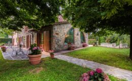 Charming stone house with large windows surrounded by lush greenery and colorful flower pots, featuring a patio area with outdoor seating and a well-maintained garden. Perfect for a serene getaway or outdoor gatherings.