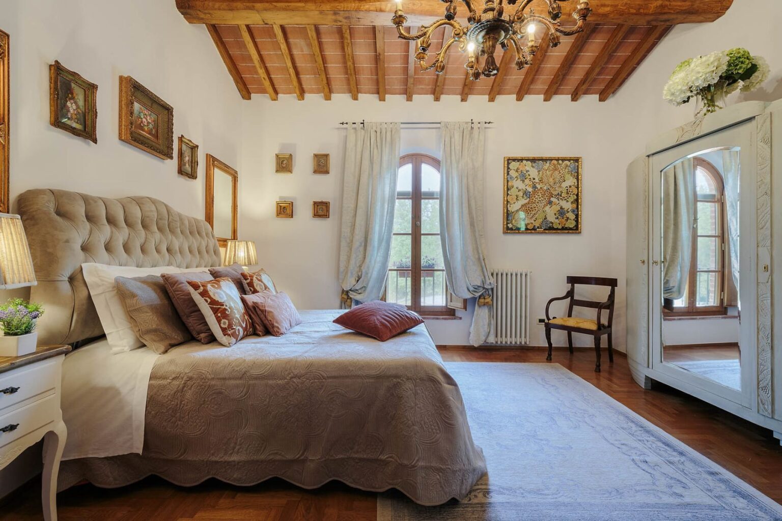 Luxurious bedroom in a villa for rent in Italy, featuring elegant decor, a plush bed with decorative pillows, wooden ceiling beams, and large windows allowing natural light. | Only on SopranoVillas