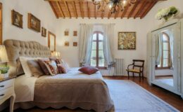 Luxurious bedroom in a villa for rent in Italy, featuring elegant decor, a plush bed with decorative pillows, wooden ceiling beams, and large windows allowing natural light.