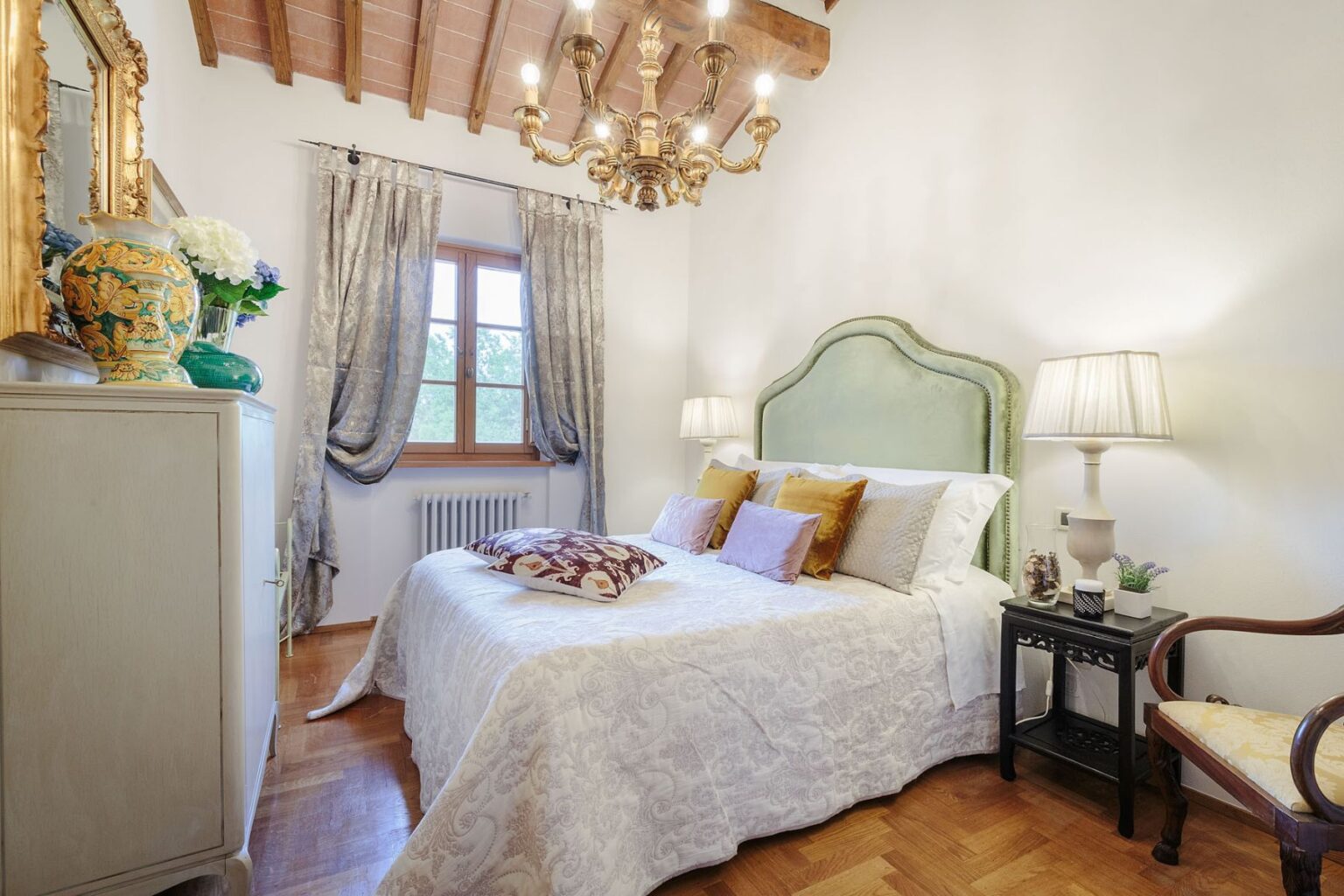 Luxury villa bedroom in Italy featuring elegant decor, a chandelier, and natural light from a window, perfect for a relaxing vacation rental experience. | Only on SopranoVillas