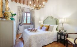 Luxury villa bedroom in Italy featuring elegant decor, a chandelier, and natural light from a window, perfect for a relaxing vacation rental experience.