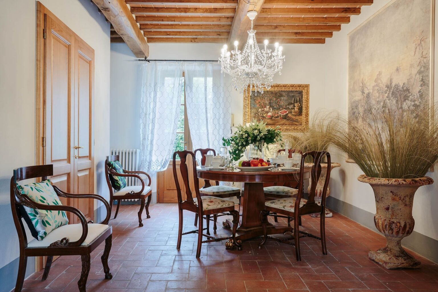 Elegant dining room featuring a wooden table surrounded by antique chairs, a crystal chandelier, and decorative plants. Sunlight filters through sheer curtains, highlighting the classic decor and warm earthy tones. | Only on SopranoVillas