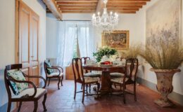 Elegant dining room featuring a wooden table surrounded by antique chairs, a crystal chandelier, and decorative plants. Sunlight filters through sheer curtains, highlighting the classic decor and warm earthy tones.