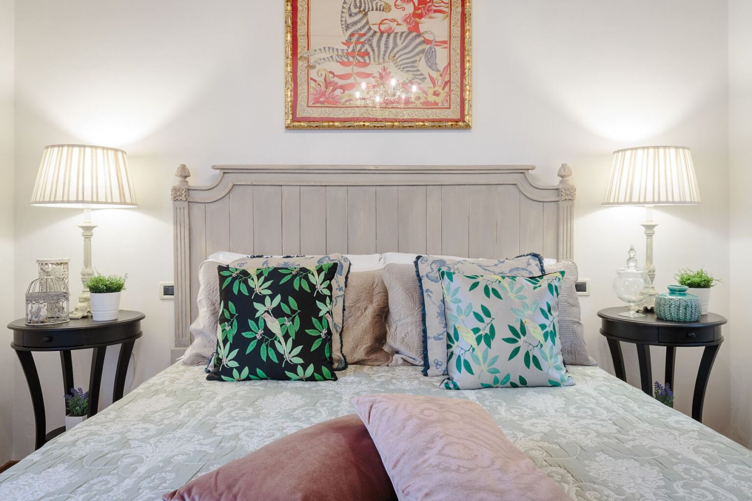 Cozy bedroom featuring a light-colored bed with decorative pillows in green and pink hues, flanked by stylish nightstands with lamps. A colorful zebra-themed artwork hangs above the bed, enhancing the room's aesthetic appeal. | Only on SopranoVillas