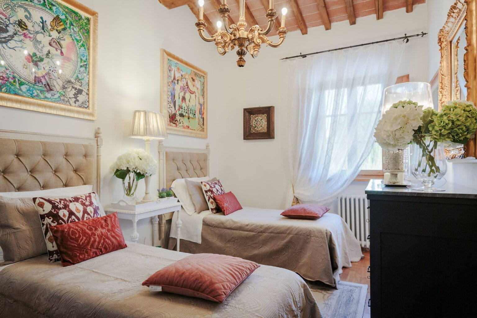 Cozy bedroom featuring two twin beds with decorative pillows, elegant chandelier, and vibrant artwork on the walls, complemented by natural light from a large window and stylish decor elements like a nightstand and floral arrangements. | Only on SopranoVillas