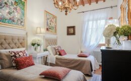 Cozy bedroom featuring two twin beds with decorative pillows, elegant chandelier, and vibrant artwork on the walls, complemented by natural light from a large window and stylish decor elements like a nightstand and floral arrangements.