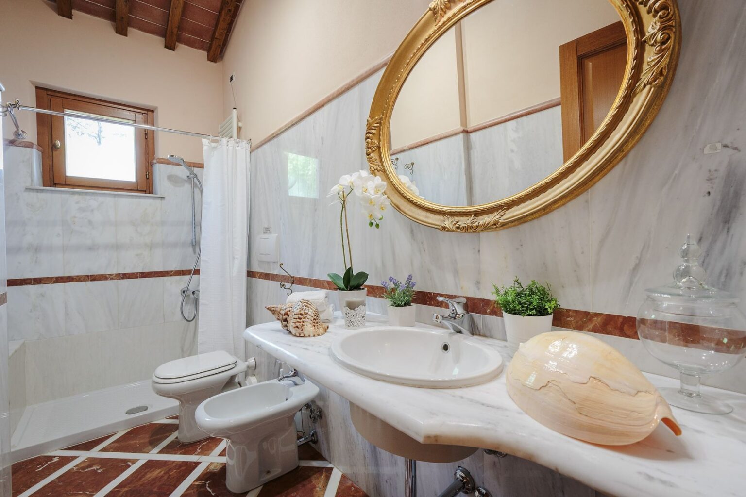 Elegant bathroom interior featuring a marble sink, gold-framed mirror, and decorative plants, with a shower area and stylish tile flooring. Ideal for showcasing modern design and luxury home features. | Only on SopranoVillas