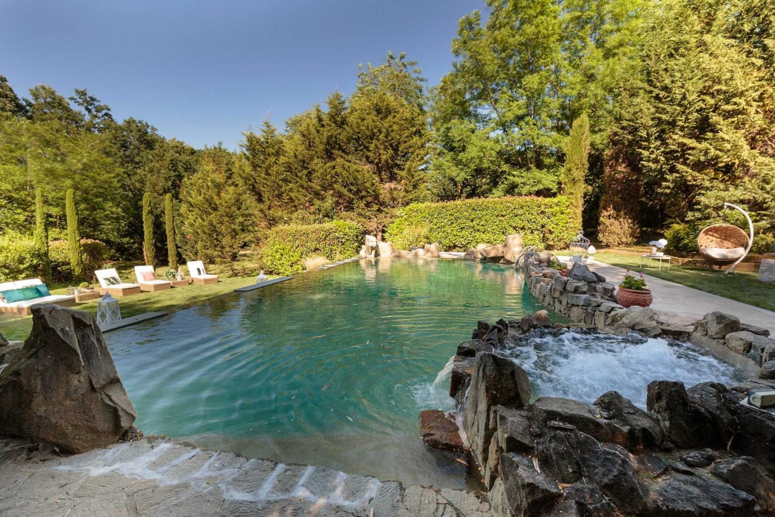 Scenic outdoor swimming pool surrounded by lush greenery, featuring natural rock formations and comfortable lounge chairs, ideal for relaxation and leisure. | Only on SopranoVillas