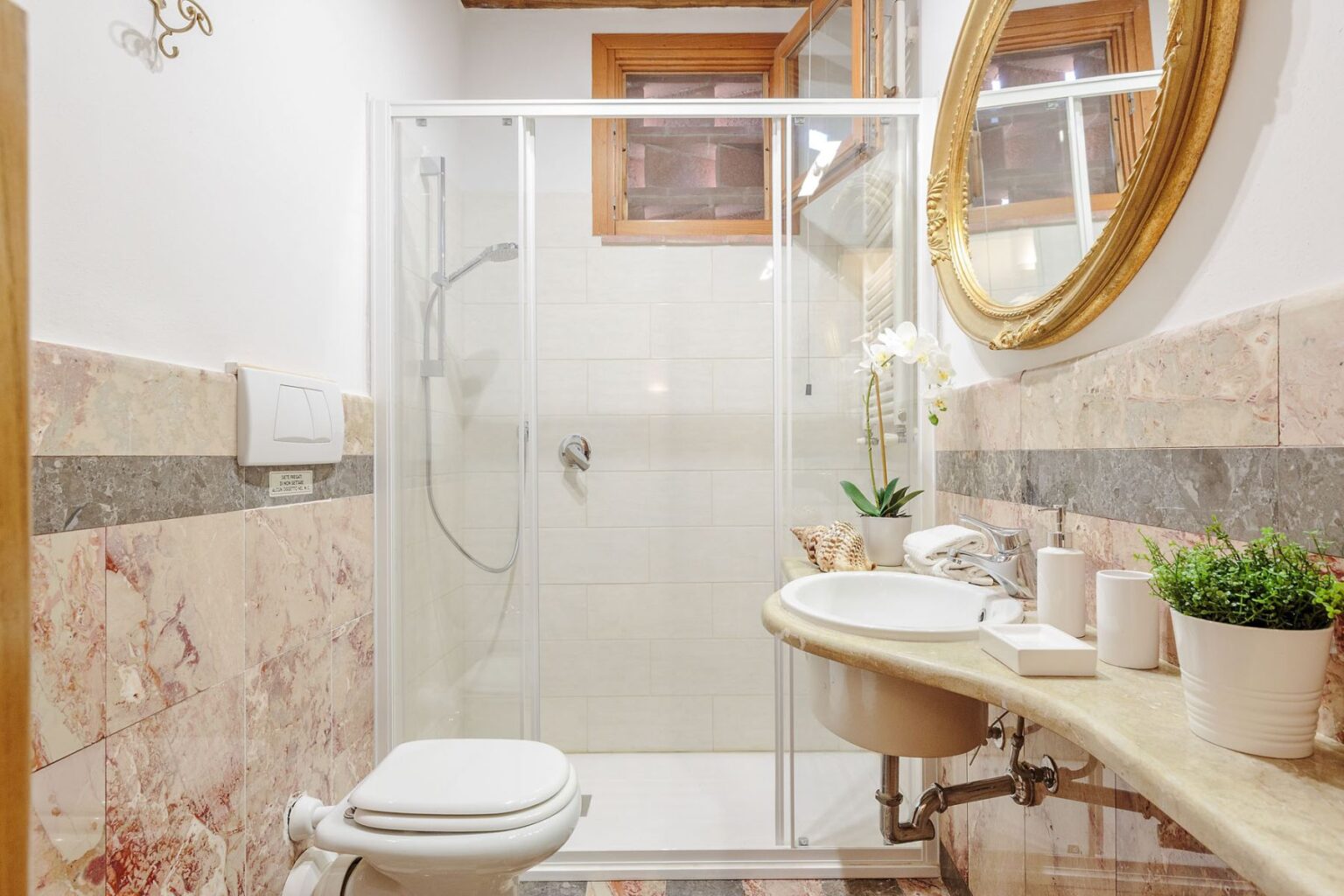 Modern bathroom interior featuring a glass shower enclosure, white toilet, and a stylish sink with a marble countertop. Natural light enters through a window, complemented by a round mirror and decorative plants. | Only on SopranoVillas
