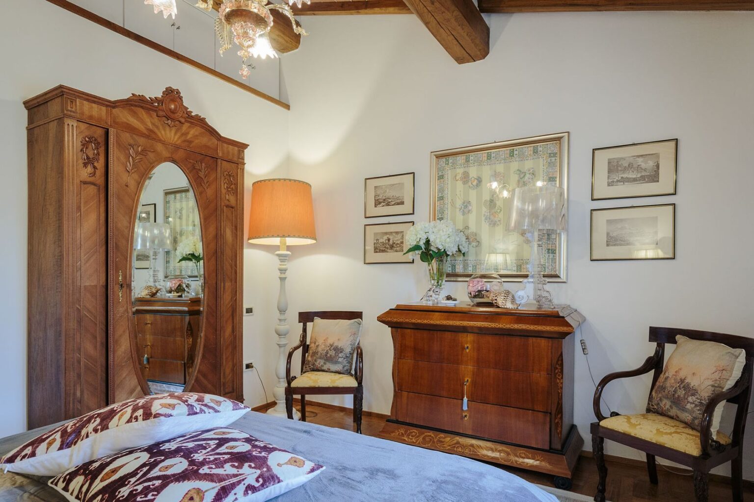 Cozy bedroom interior featuring a vintage wooden wardrobe with a mirror, an elegant wooden chest of drawers, two stylish chairs, and framed artwork on the walls, illuminated by a warm lamp. | Only on SopranoVillas