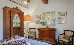 Cozy bedroom interior featuring a vintage wooden wardrobe with a mirror, an elegant wooden chest of drawers, two stylish chairs, and framed artwork on the walls, illuminated by a warm lamp.
