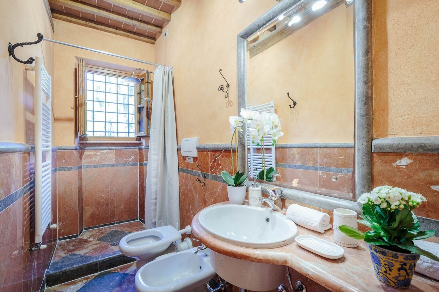 Cozy bathroom interior featuring a shower, sink, and toilet, with warm orange walls, decorative plants, and a large mirror. Ideal for home renovation inspiration. | Only on SopranoVillas