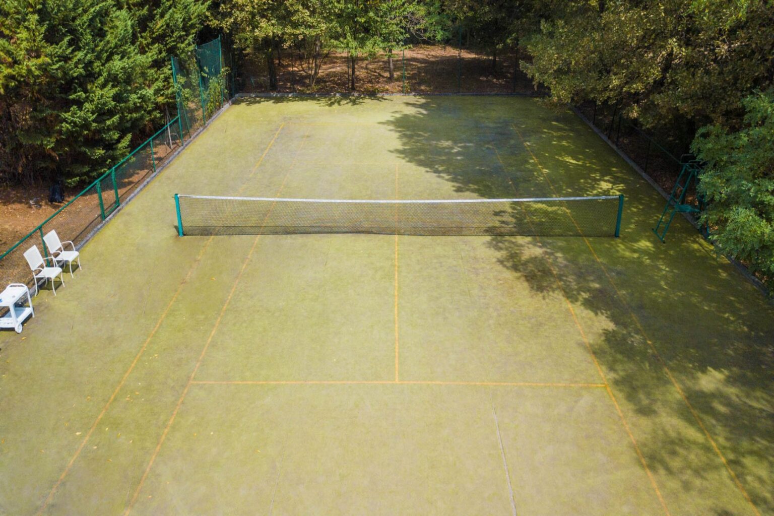 Aerial view of a tennis court with a net in the center, surrounded by trees and overgrown grass, featuring two white chairs nearby. | Only on SopranoVillas