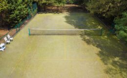 Aerial view of a tennis court with a net in the center, surrounded by trees and overgrown grass, featuring two white chairs nearby.