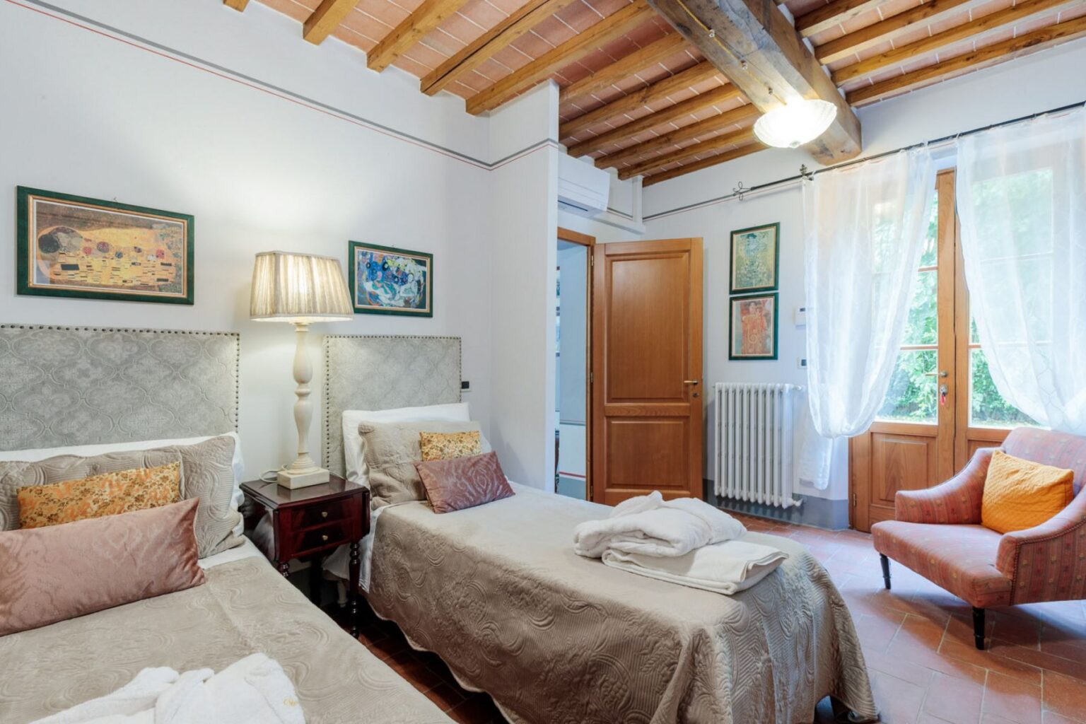 Cozy guest room with two twin beds, warm wooden beams, a decorative lamp, and natural light streaming through large windows, featuring soft furnishings and a comfortable armchair. Ideal for relaxation and comfort in a stylish setting. | Only on SopranoVillas