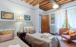 Cozy guest room with two twin beds, warm wooden beams, a decorative lamp, and natural light streaming through large windows, featuring soft furnishings and a comfortable armchair. Ideal for relaxation and comfort in a stylish setting.
