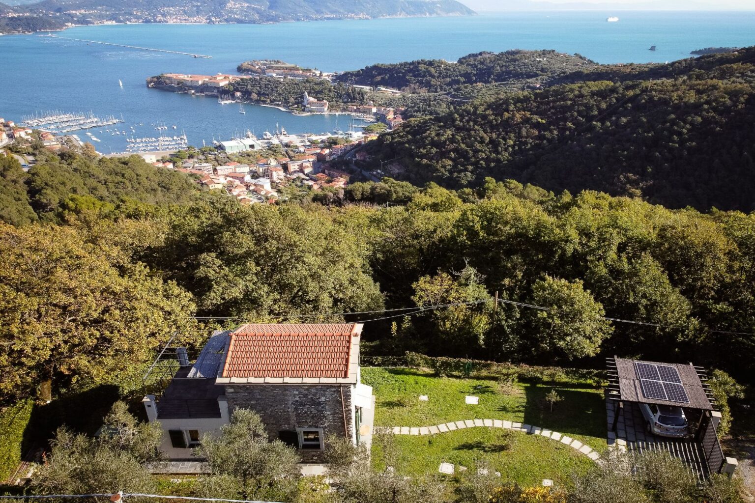 Aerial view of a modern house with a red-tiled roof surrounded by lush greenery, overlooking a picturesque coastal town and marina with boats in the water. The property features a solar panel carport and a landscaped garden with curved pathways. | Only on SopranoVillas