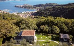 Aerial view of a modern house with a red-tiled roof surrounded by lush greenery, overlooking a picturesque coastal town and marina with boats in the water. The property features a solar panel carport and a landscaped garden with curved pathways.