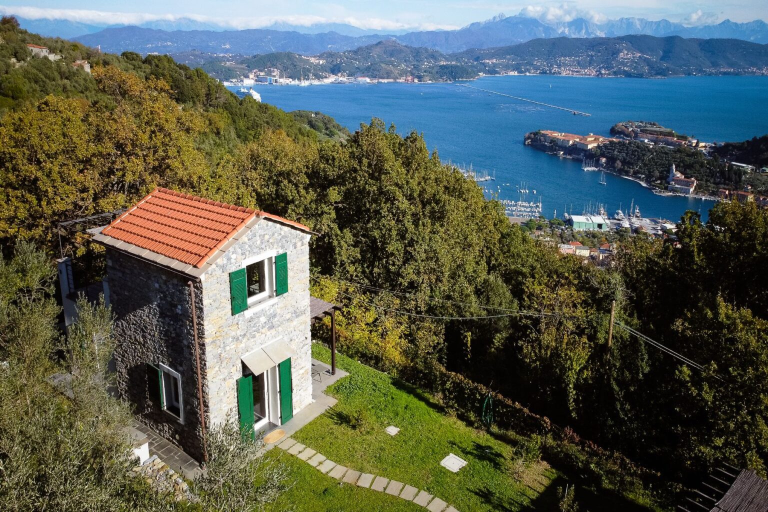 Scenic view of a charming stone house with green shutters, surrounded by lush greenery and overlooking a picturesque bay with boats and mountains in the background. Ideal for nature lovers and travelers seeking tranquility. | Only on SopranoVillas