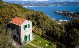 Scenic view of a charming stone house with green shutters, surrounded by lush greenery and overlooking a picturesque bay with boats and mountains in the background. Ideal for nature lovers and travelers seeking tranquility.
