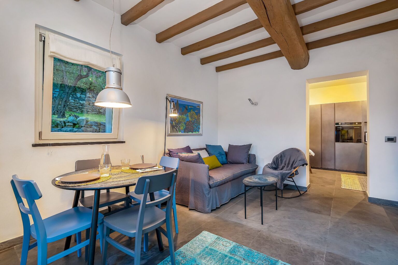 Cozy living and dining area featuring a round table with blue chairs, a comfortable sofa adorned with colorful cushions, and a modern kitchen visible in the background. Natural light filters through a window, highlighting the wooden beams and stylish decor. Perfect for relaxation and entertaining. | Only on SopranoVillas