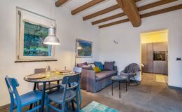 Cozy living and dining area featuring a round table with blue chairs, a comfortable sofa adorned with colorful cushions, and a modern kitchen visible in the background. Natural light filters through a window, highlighting the wooden beams and stylish decor. Perfect for relaxation and entertaining.