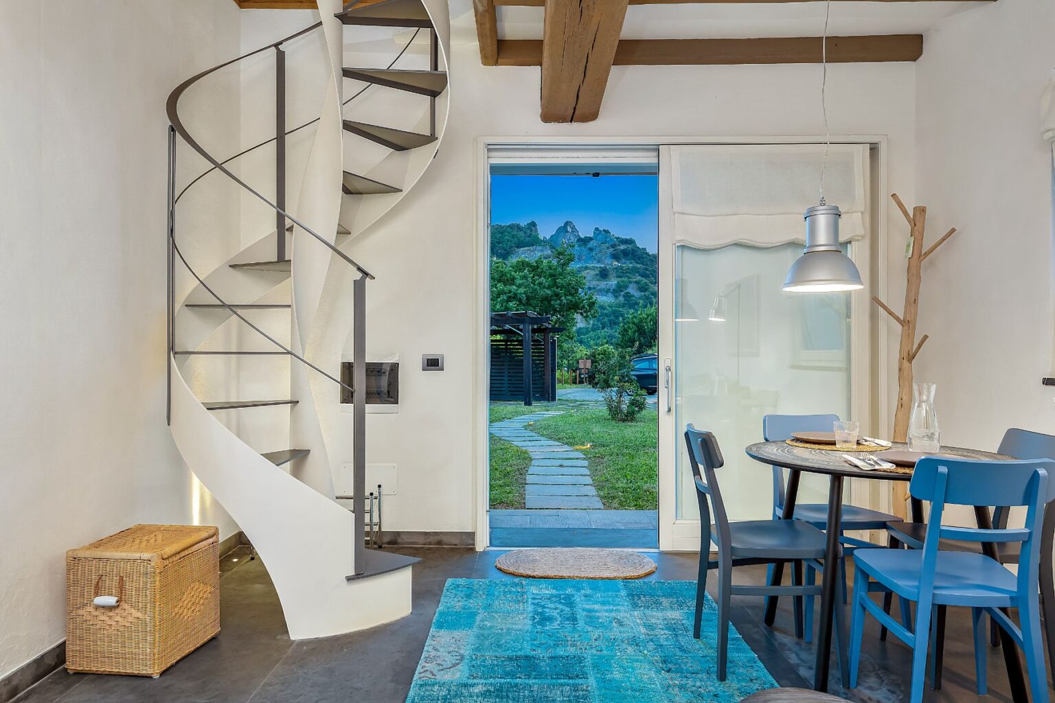 Modern interior design featuring a spiral staircase, a cozy dining area with a round table and blue chairs, and a view of a garden through glass doors. The space is illuminated with natural light, showcasing a stylish rug and a wicker storage box. | Only on SopranoVillas