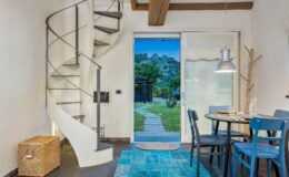 Modern interior design featuring a spiral staircase, a cozy dining area with a round table and blue chairs, and a view of a garden through glass doors. The space is illuminated with natural light, showcasing a stylish rug and a wicker storage box.