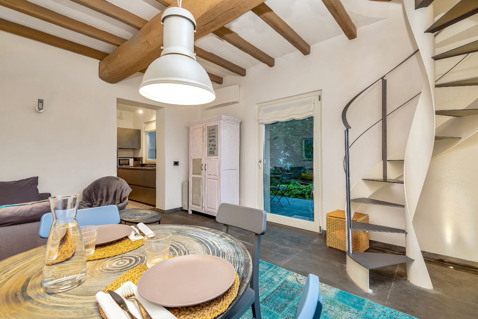 Cozy living space featuring a modern dining area with a circular table, stylish decor, and a spiral staircase. The room has wooden beams, a soft sofa, and views of an outdoor patio through a glass door, creating a welcoming atmosphere. | Only on SopranoVillas