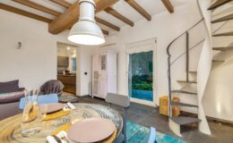 Cozy living space featuring a modern dining area with a circular table, stylish decor, and a spiral staircase. The room has wooden beams, a soft sofa, and views of an outdoor patio through a glass door, creating a welcoming atmosphere.