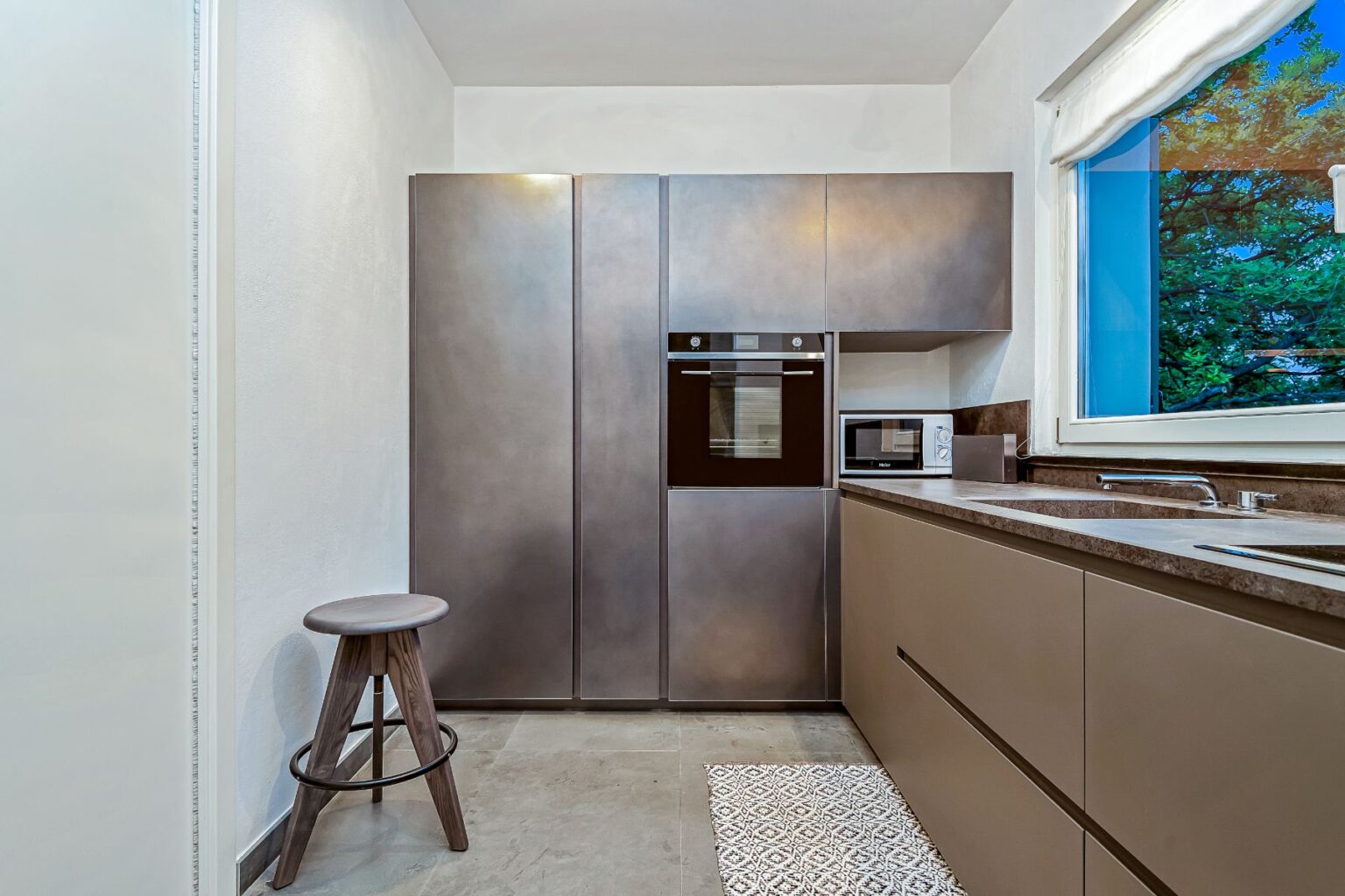 Modern kitchen interior featuring sleek, metallic cabinetry, built-in oven, and microwave. A wooden stool complements the minimalist design, with a window showcasing greenery outside. Ideal for contemporary home decor inspiration. | Only on SopranoVillas