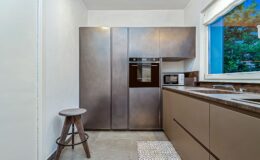 Modern kitchen interior featuring sleek, metallic cabinetry, built-in oven, and microwave. A wooden stool complements the minimalist design, with a window showcasing greenery outside. Ideal for contemporary home decor inspiration.