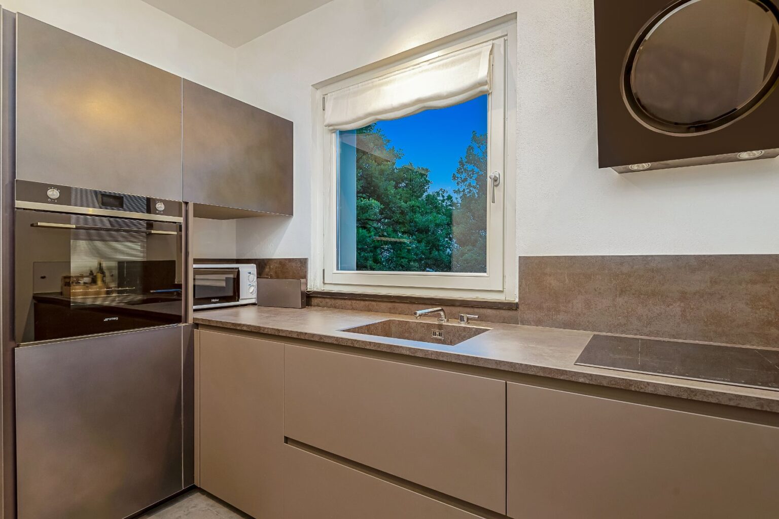 Modern kitchen interior featuring sleek cabinetry, stainless steel appliances, and a large window with a view of greenery. The design includes a built-in oven, microwave, and a minimalist sink area, emphasizing functionality and style in contemporary home design. | Only on SopranoVillas