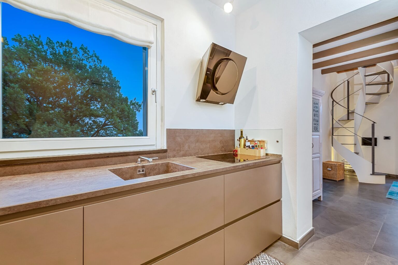 Modern kitchen interior featuring a sleek countertop with a double sink, under-cabinet lighting, and a stylish extractor hood. A large window offers a view of a lush tree, enhancing the natural light in the space. A spiral staircase is visible in the background, adding an elegant design element. | Only on SopranoVillas
