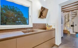 Modern kitchen interior featuring a sleek countertop with a double sink, under-cabinet lighting, and a stylish extractor hood. A large window offers a view of a lush tree, enhancing the natural light in the space. A spiral staircase is visible in the background, adding an elegant design element.