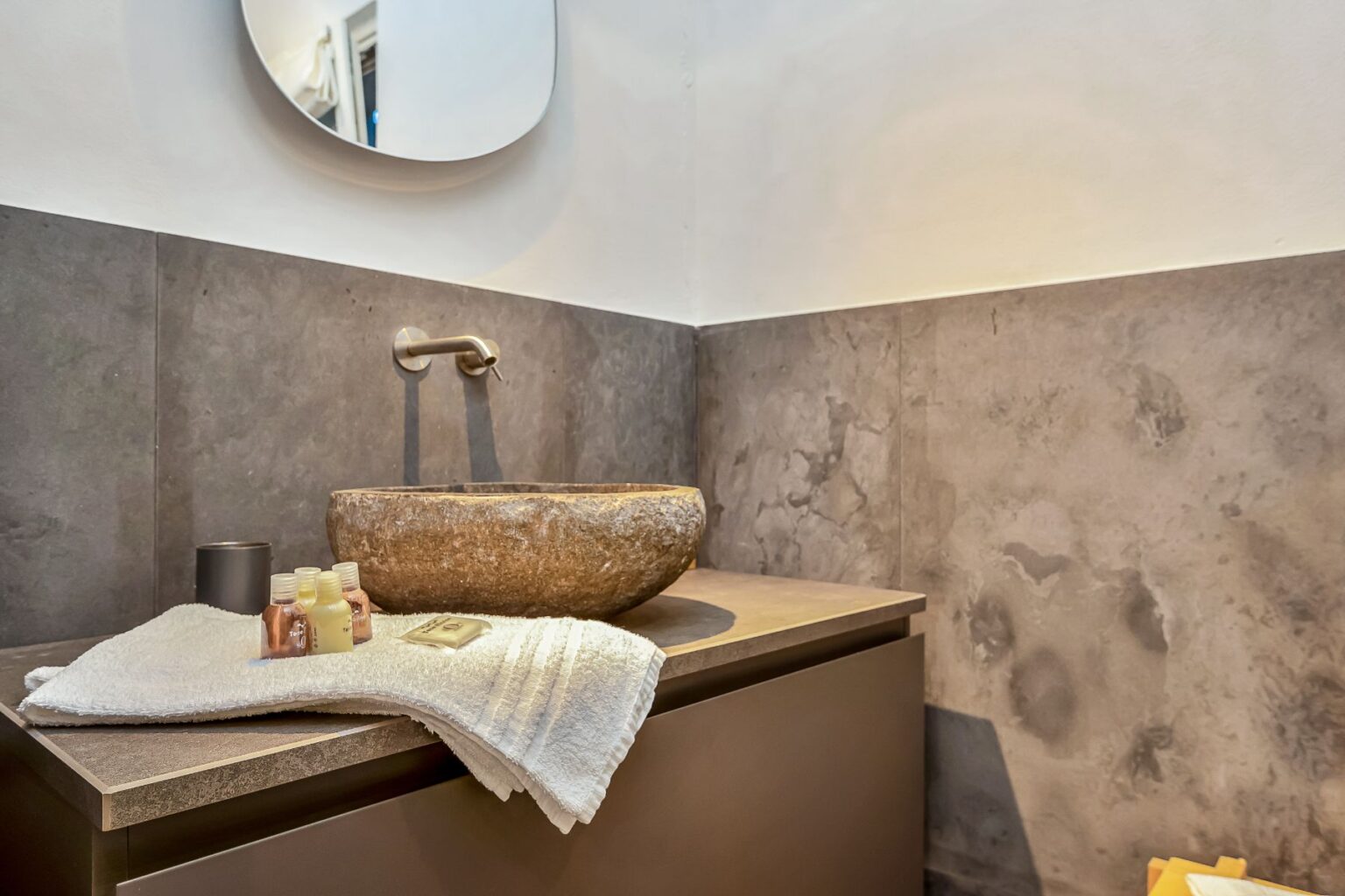 Modern bathroom interior featuring a stone sink, sleek faucet, and decorative toiletries on a white towel. The wall is adorned with textured gray tiles, and a round mirror reflects the space, enhancing the contemporary design. | Only on SopranoVillas
