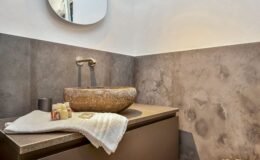 Modern bathroom interior featuring a stone sink, sleek faucet, and decorative toiletries on a white towel. The wall is adorned with textured gray tiles, and a round mirror reflects the space, enhancing the contemporary design.
