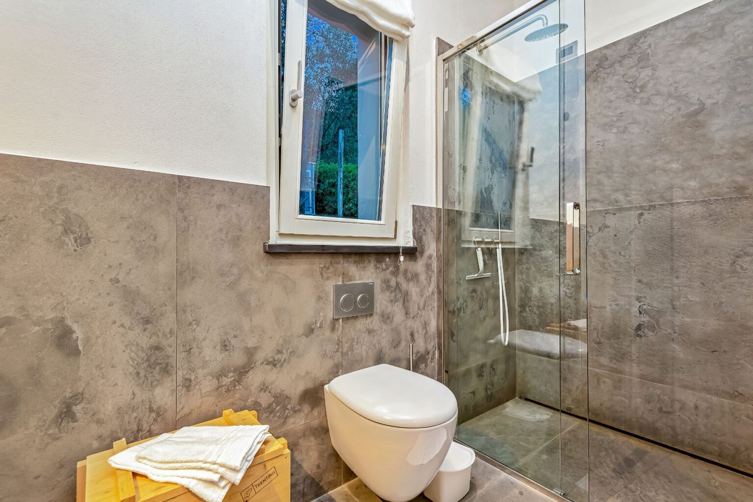 Modern bathroom featuring a glass shower, sleek toilet, and stylish gray stone walls, with natural light coming through a window. | Only on SopranoVillas