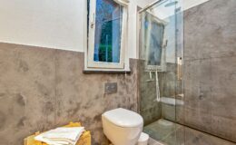 Modern bathroom featuring a glass shower, sleek toilet, and stylish gray stone walls, with natural light coming through a window.